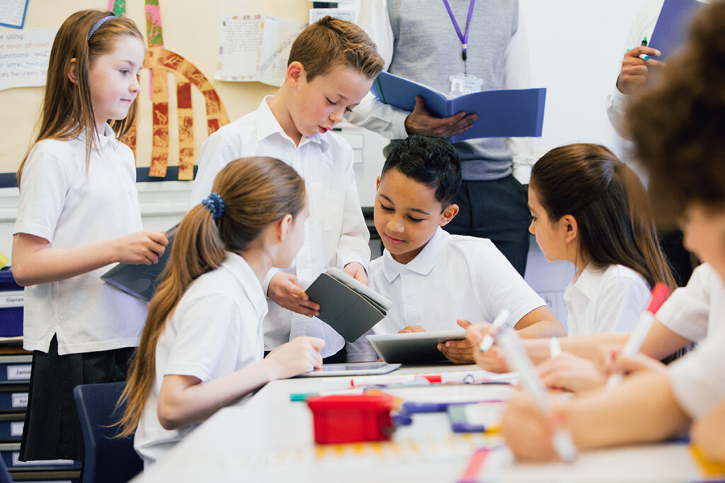 A group of school children can be seen working on digital tablets and whiteboards, they are all working happily. Two unrecognisable teachers can be seen in the background.