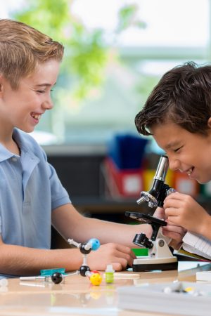 Caucasian and Hispanic elementary private school boys use a microscope in science class. They are working on a project together. The Hispanic boy looks into the microscope while the Caucasian boy watches. They are wearing school uniforms.
